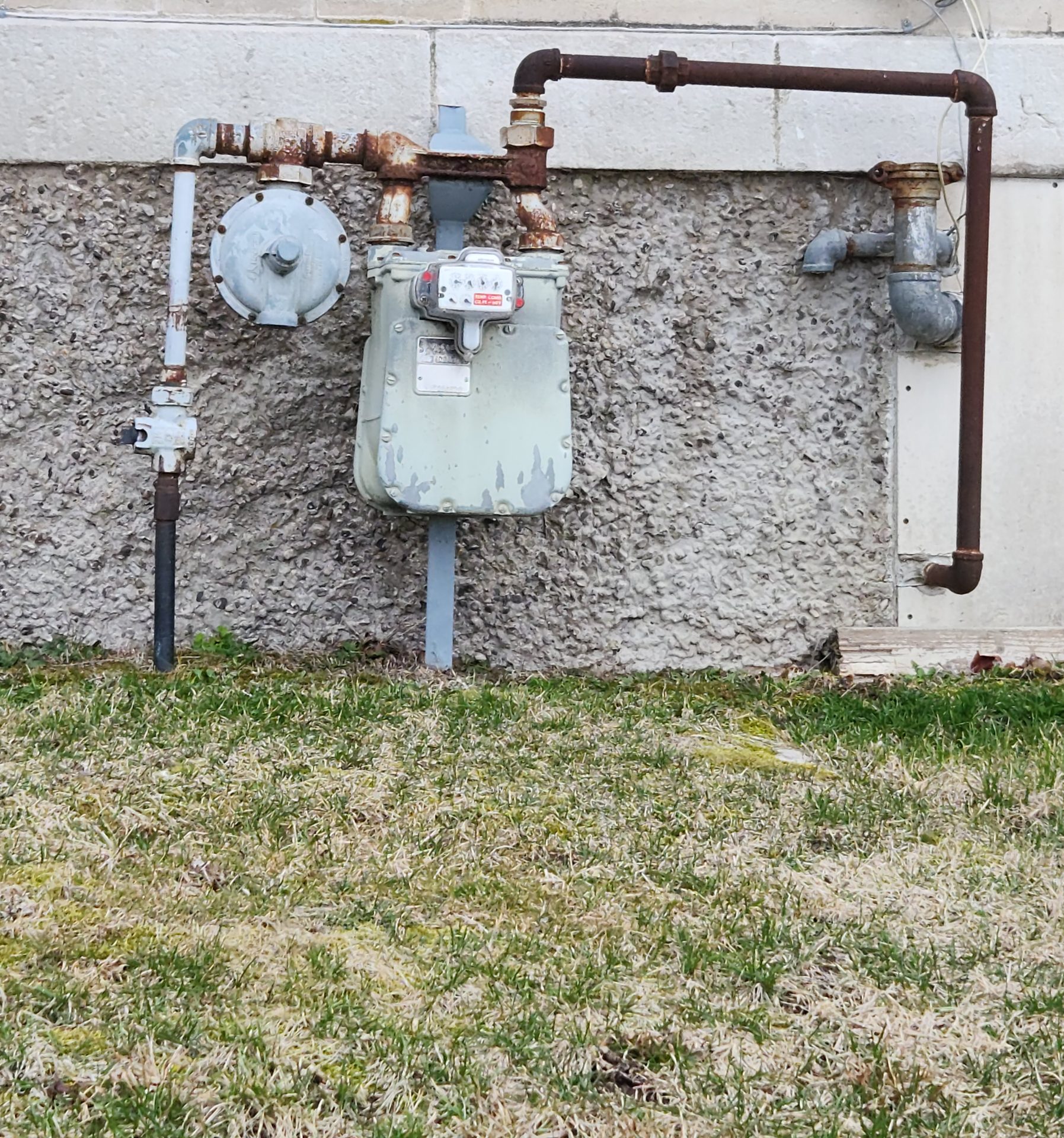 An outdoor gas meter with rusty black steel pipe running up the wall and into the home, showing the point where the entire home's gas supply begins.
